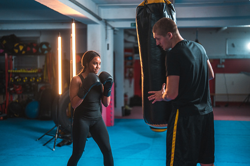 Strong young multiracial female athlete punching a boxing bag. She is indoors, training with her boxing coach.