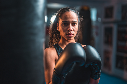 Portrait of a confident young multiracial female boxer getting ready for a fight. She is indoors in a gym, wearing boxing gloves.