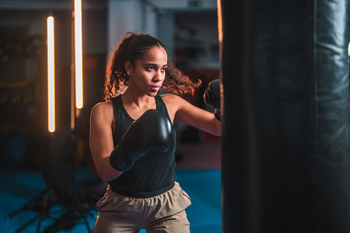 Attractive young Latin American female athlete punching a boxing bag with boxing gloves. She is indoors in a gym, focused on her training.