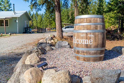 A rustic barrel with a welcome message alongside a gravel driveway, outside a rural home on acreage with a wooded lot in North Idaho, USA.