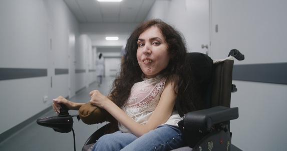 Woman with spinal muscular atrophy in motorized wheelchair in the middle of clinic corridor smiles and looks at camera. Medical staff walk in hallway of hospital or medical facility in the background.