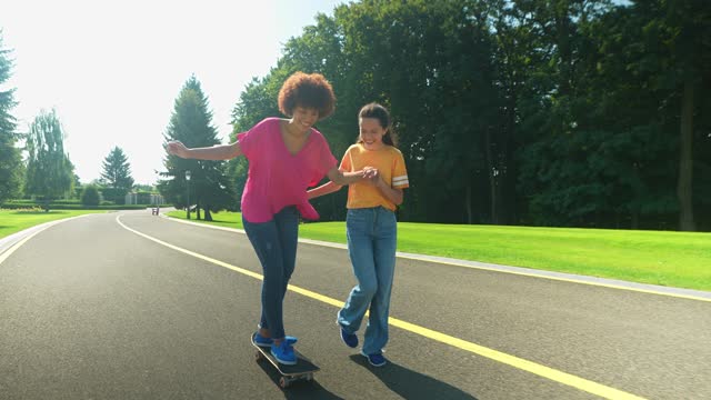 Cheerful African American mother skateboarding with help of daughter outdoor