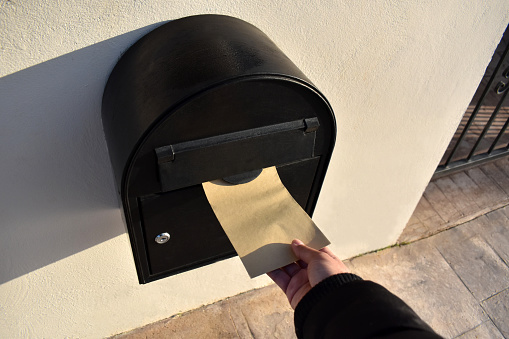 Hand putting a letter or a brown envelope in the mailbox in front of a house with copy space on the right side and on the envelope.