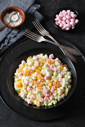 Ambrosia Salad with pineapple, mandarin oranges, yogurt, mini marshmallows, coconut and whipped cream in black bowl on concrete table, american recipe, vertical view