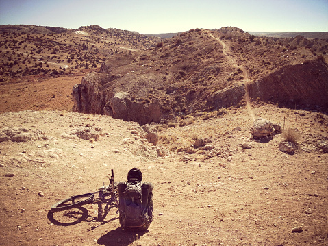 a mountain biker man takes a break and rests sitting down next to his bicycle with the trail and desert badlands landscape beyond.  such beautiful nature scenery and outdoor sports and adventure can be found at the white mesa bike trails, san ysidro, new mexico.  horizontal wide angle and high angle mobilestock composition with retro color treatment.  