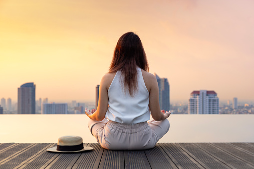 Back of woman relaxingly practicing meditation at the swimming pool rooftop with view of urban skyline building to attain happiness from inner peace wisdom