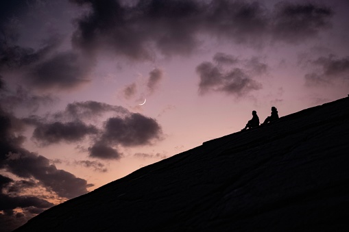 On a rocky hill, two people’s silhouette embraces the enchanting crescent moon, crafting a picturesque tableau that captures the essence of romance and natural beauty.
