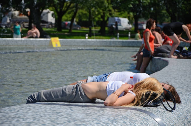 Warsaw, Poland - May 22nd, 2012: Two young girls: a blonde and a brunette, lying and sunbathing on a fountain border. Other people sitting on the border and standing in the water on the right. Park trees in the background