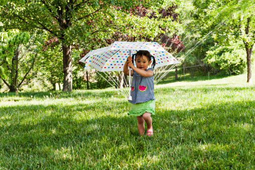 LIttle girl holding an umbrella under a sprinkler to keep from getting wet.