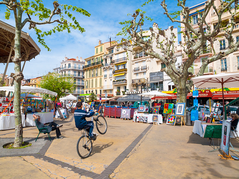 Cannes, France - April 24, 2010: Small local market in the square in Cannes. People can be seen sitting at their stalls, while a youth passes doing a stunt on his bike.
