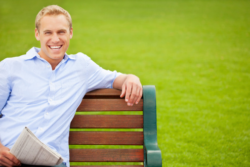 Portrait of a cheerful young guy sitting on a park bench with newspaper. Horizontal shot.