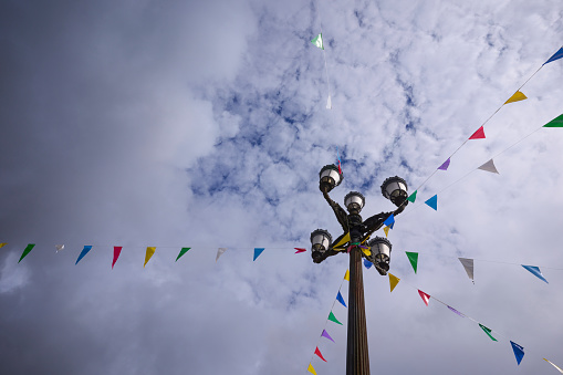 Diagonal lines of colored pennants fluttering in the wind, hanging from an old street light, over a cloudy sky. Summer popular party background with copy space.