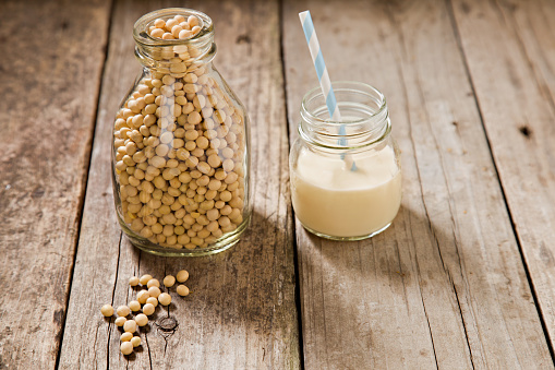A close up shot of an old milk bottle filled with dry soy beans with a few beans on the foreground and a small glass jar container with soy milk and a straw. Shot on an old grungy wooden table. 