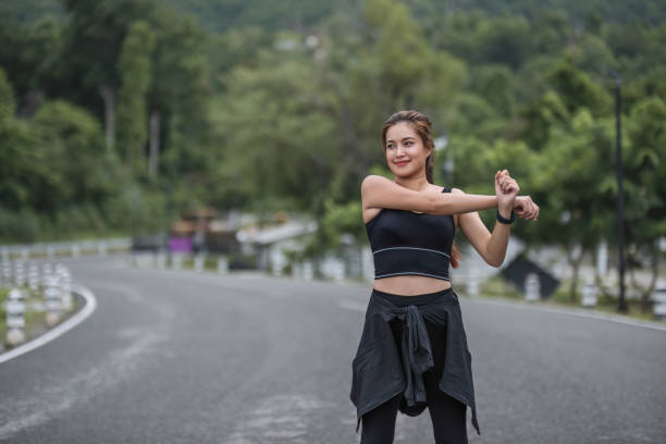 um retrato de uma jovem asiática esportiva e feliz em roupas esportivas está esticando os braços, aquecendo seu corpo antes de correr ao ar livre. conceito de atividade ao ar livre - aquecimento físico - fotografias e filmes do acervo