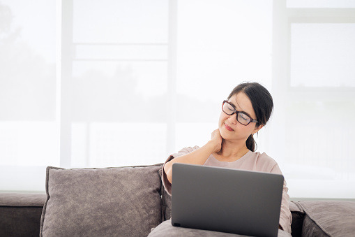 Mature middle age asian woman feeling pain in neck and shoulder after working on computer laptop for a long time in living room at home . She stretches to relax her muscles.Office syndrome concept.