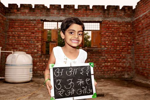 Poor elementary age school girl holding chalkboard or slate in hands with Hindi alphabets writing on it portrait against brick wall.