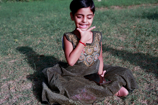 Cheerful cute little girl of Indian ethnicity sitting on the grass in the park and using mobile phone portrait close up