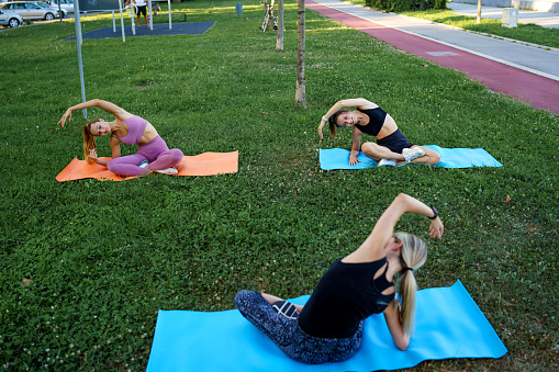 Three young Caucasian women having a yoga class at the park. They're sitting on yoga mats and stretching.
