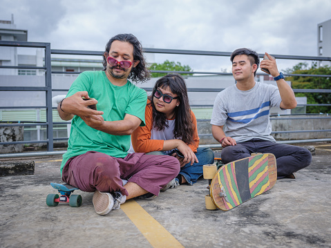 Small Group of young Asian skateboarder man take a selfie with mobile phone after play skateboard at rooftop parking outdoors, Active Lifestyle