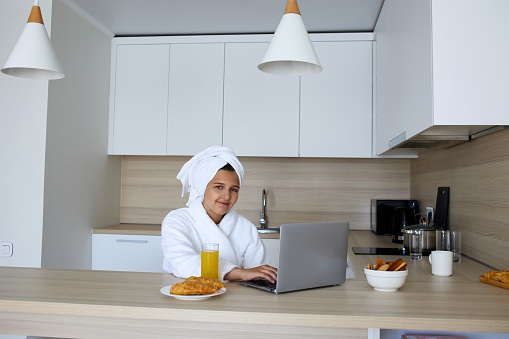 A young girl in a bathrobe is working at a laptop in the kitchen. In front of her is a glass of orange juice and pastries. Breakfast time.