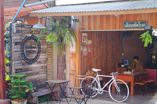Small coffee shop in Chiang Khan in Loei province. Old wooden house. A bicycle is placed outside. A man is sitting inside and is using laptop. View from street into open shop.
