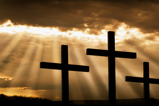 Dramatic sky silhouettes three wooden crosses with shafts of sunlight breaking through the clouds. A dramatic and inspiring religious photographic illustration for Christian beliefs including Easter and Good Friday.