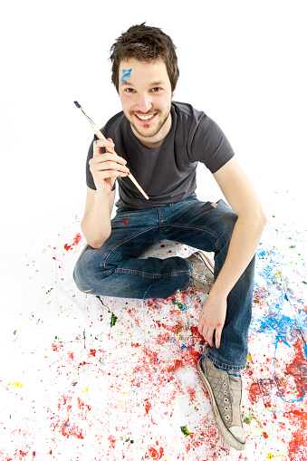 cheerful young painter with brush sitting on color spots, isolated on white. 