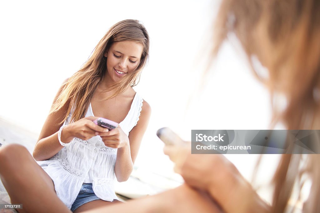 Due ragazze adolescenti felici i messaggi di testo sulla spiaggia - Foto stock royalty-free di 18-19 anni