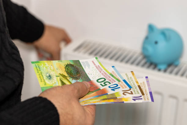 frais de chauffage des appartements en hiver en suisse, concept énergétique et économique, homme allumant le radiateur, tenant une pile de francs suisses dans sa main et une tirelire debout sur le radiateur - switzerland swiss currency chf euro symbol photos et images de collection