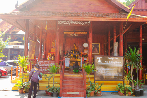 Praying thai woman at Sri Khun Mueang in Chiang Khan in morning sunshine. Woman is praying at a small wooden shrine. Inside of shrine are statues, a statue of a monk and several images. A person is watering plants in front of the shrine. Chiang Khan is in Loie province