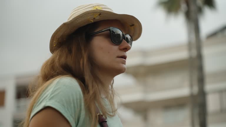 A young woman wearing a straw hat and sunglasses strolls through the city, with palm trees in the background. The view is from a low angle, and the scene is in slow motion.