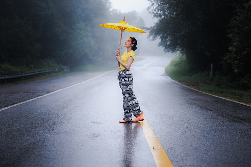 portrait of young woman standing and holding umblella in the middle of the road in the rainy season, natural forest and concept.