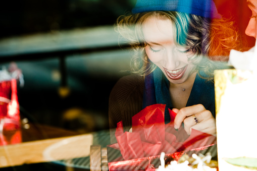 Shot of Pretty blond girl wearing a knit hat and sweater viewed through a window smiling opening a gift with red wrapping paper signifying perhaps a Christmas or Valentine's Day gift.  Window glare gives image a nice authentic and candid feel.