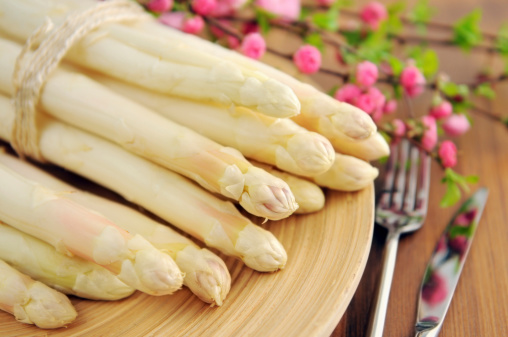 Bundle fresh white Asparagus on cup with pink blooming almond in background. Aside laying table knife and fork.