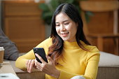 happy smiling asian young woman in yellow sweater sitting on sofa and having video call on smartphone at home