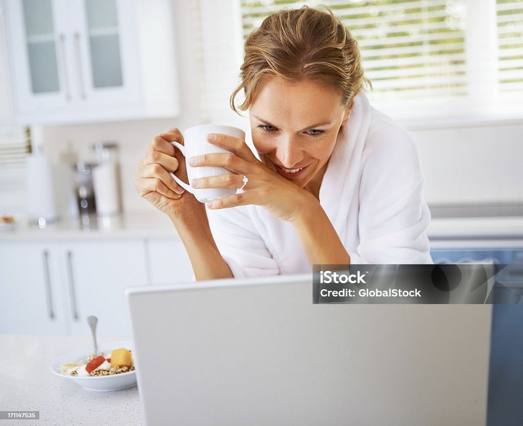 Młoda kobieta Trzymając kubek kawy i Patrząc na laptopa - Zbiór zdjęć royalty-free (Jedzenie)