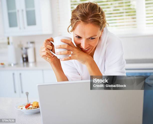 Młoda Kobieta Trzymając Kubek Kawy I Patrząc Na Laptopa - zdjęcia stockowe i więcej obrazów Jedzenie
