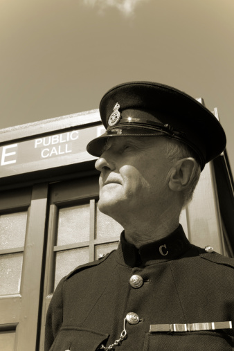 A world war 2 era british bobby stands by his Police box, his cap sheltering his face from the sun