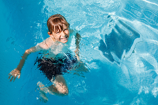Portrait of happy little kid boy in the pool and having fun on family vacations in a hotel resort. Healthy child playing in water, swimming and splashing