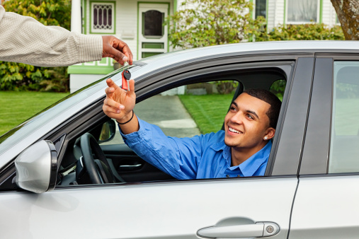 Photo of a young man behind the wheel of a silver sports car, taking the keys from his friend with a smile.