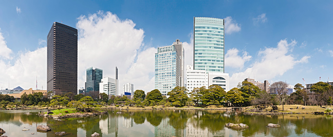 Big blue panoramic spring skies and white fluffy clouds over the skyscrapers and high rises, landmarks and downtown architecture of Shiodome and Shimbashi reflecting in the tranquil waters of the Sensui pond in the urban green oasis of Kyo Shiba Rikyo Onshi Teien, Minato-Ku, central Tokyo, Japan. ProPhoto RGB profile for maximum color fidelity and gamut.
