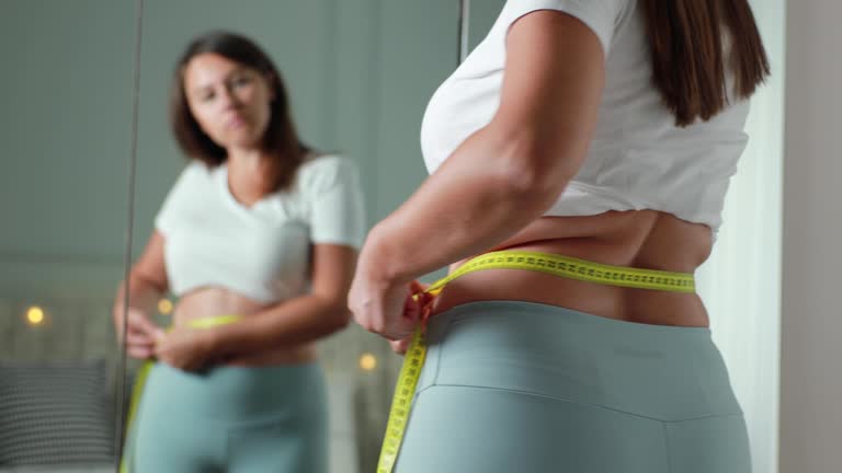 Young woman taking measurements of her body in front of the mirror in her bedroom