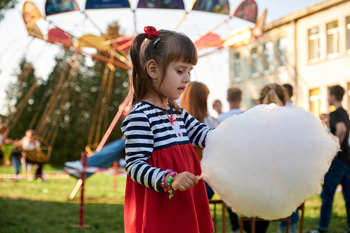 Cute little girl eating sweet cotton wool against the backdrop of rides, carousels. Close-up portrait with sweet cotton. A four-year-old girl is eating cotton candy in the park.