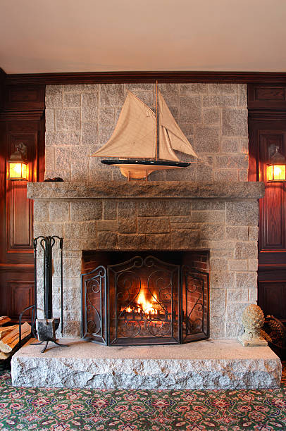 A stone fireplace with a cozy burning fire.  A sail boat model sits on top of the mantel piece.  Walls are wood paneled and there is a carpet on the floor.