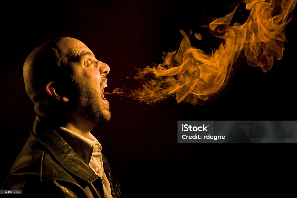 Man Breathing Fire - Heartburn, Bad Breath, or Anger Bald, Caucasian man in his 30s breathing fire from his mouth. His face is lit from the glow of the flames. Concept shot for heartburn, bad breath, or anger. Fire-eater Stock Photo