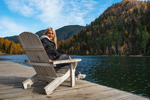 A Woman sits in an Adirondack chair on a wooden dock overlooking the blue water of the Lake. Blonde woman traveler tourist in sportswear admires the view of the  lake, mountains, autumn