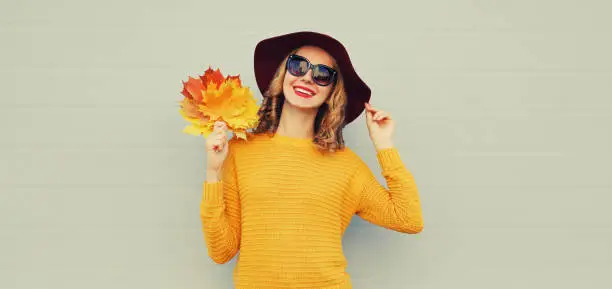 Autumn portrait of happy smiling young woman holds yellow maple leaves wearing round hat, sweater on gray background
