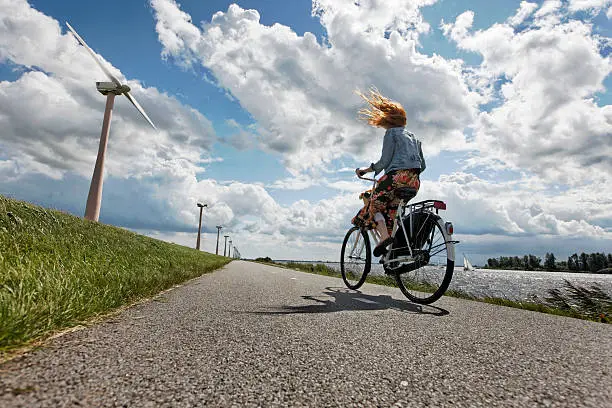 young woman on her bicycle traveling on a wind blown dike in Holland, wind turbines in the background