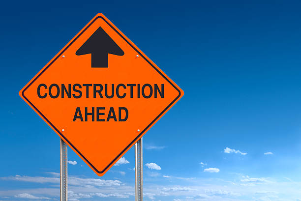 Orange Warning Construction Ahead Message Road Sign Over Blue Sky Conceptual road sign warning of construction ahead - a clipping path is included to separate sign from bkg. construction zone signs stock pictures, royalty-free photos & images