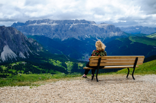 An older woman sitting on a bench overlooking dramatic mountain scenery and clouds. Location: Mt. Seceda, Val Gardena, Italy
